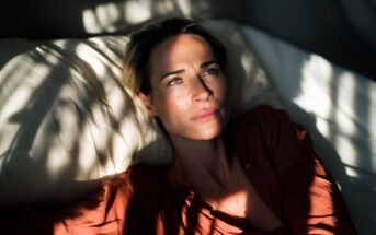 A woman with short blonde hair lies on a pillow in sunlight, wearing a rust-colored shirt, looking thoughtful as shadows from leaves fall across her face and the bedding.
