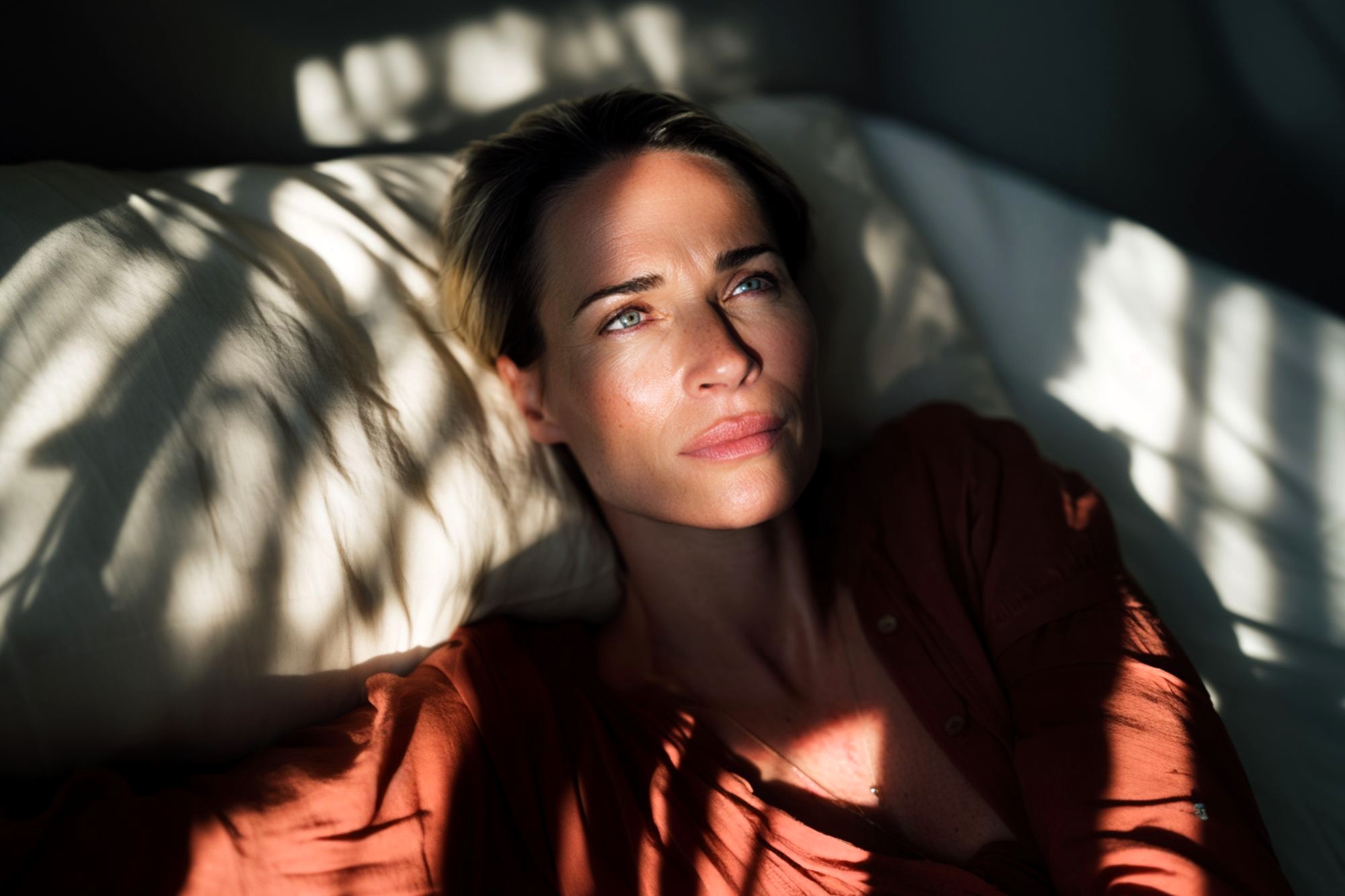 A woman with short blonde hair lies on a pillow in sunlight, wearing a rust-colored shirt, looking thoughtful as shadows from leaves fall across her face and the bedding.