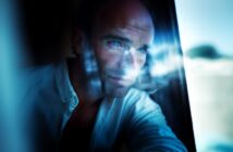 A man with blue eyes stares thoughtfully out of a window, with soft natural light and reflections illuminating his face.