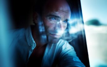 A man with blue eyes stares thoughtfully out of a window, with soft natural light and reflections illuminating his face.