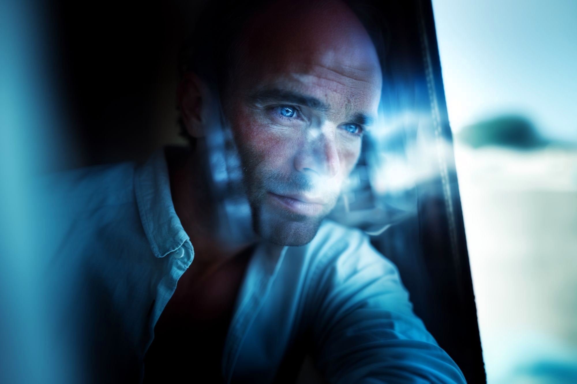 A man with blue eyes stares thoughtfully out of a window, with soft natural light and reflections illuminating his face.