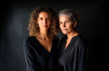 Two women, one younger with curly hair and one older with gray curly hair, stand close together against a dark background, both wearing black tops and looking seriously at the camera.
