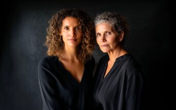 Two women, one younger with curly hair and one older with gray curly hair, stand close together against a dark background, both wearing black tops and looking seriously at the camera.
