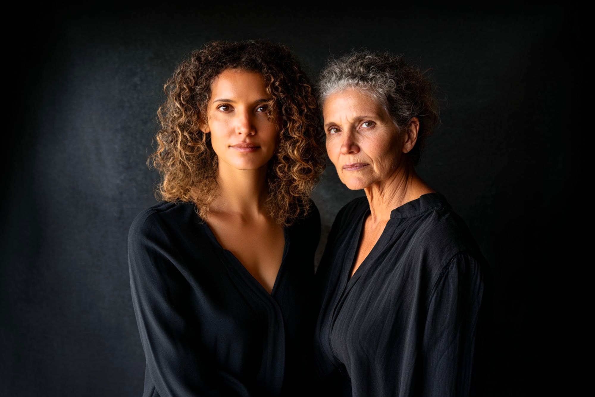 Two women, one younger with curly hair and one older with gray curly hair, stand close together against a dark background, both wearing black tops and looking seriously at the camera.