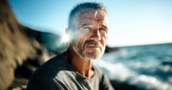 A man with gray hair and a beard sits by the ocean, sunlight shining behind him. He wears a gray shirt and looks calmly at the camera, with waves and rocky cliffs in the background.