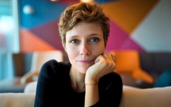 A woman with short, wavy hair and a gentle smile sits indoors, resting her chin on her hand. The background features colorful geometric wall patterns and blurred furniture.