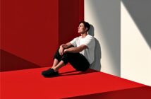 A man in a white shirt and black pants sits on a red floor, leaning against a wall with sunlight casting shadows. The setting is minimalist, with bold red and white geometric surfaces.