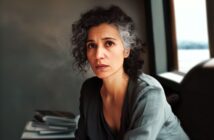 A woman with curly gray hair and a serious expression sits indoors near a window, wearing a loose gray top. A stack of magazines or books is blurred in the background.