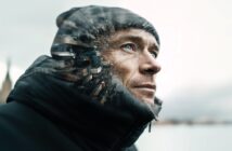A man in a winter jacket and beanie looks upward. The left side of his face blends into a cityscape, creating a double exposure effect. The background is blurred and light, suggesting a cold outdoor setting.