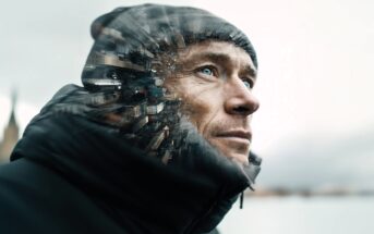 A man in a winter jacket and beanie looks upward. The left side of his face blends into a cityscape, creating a double exposure effect. The background is blurred and light, suggesting a cold outdoor setting.