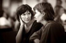 Two women sit close together, having an intimate conversation. One woman listens attentively with her hand to her face, while the other speaks. The background is softly blurred, focusing attention on their expressions.