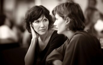 Two women sit close together, having an intimate conversation. One woman listens attentively with her hand to her face, while the other speaks. The background is softly blurred, focusing attention on their expressions.