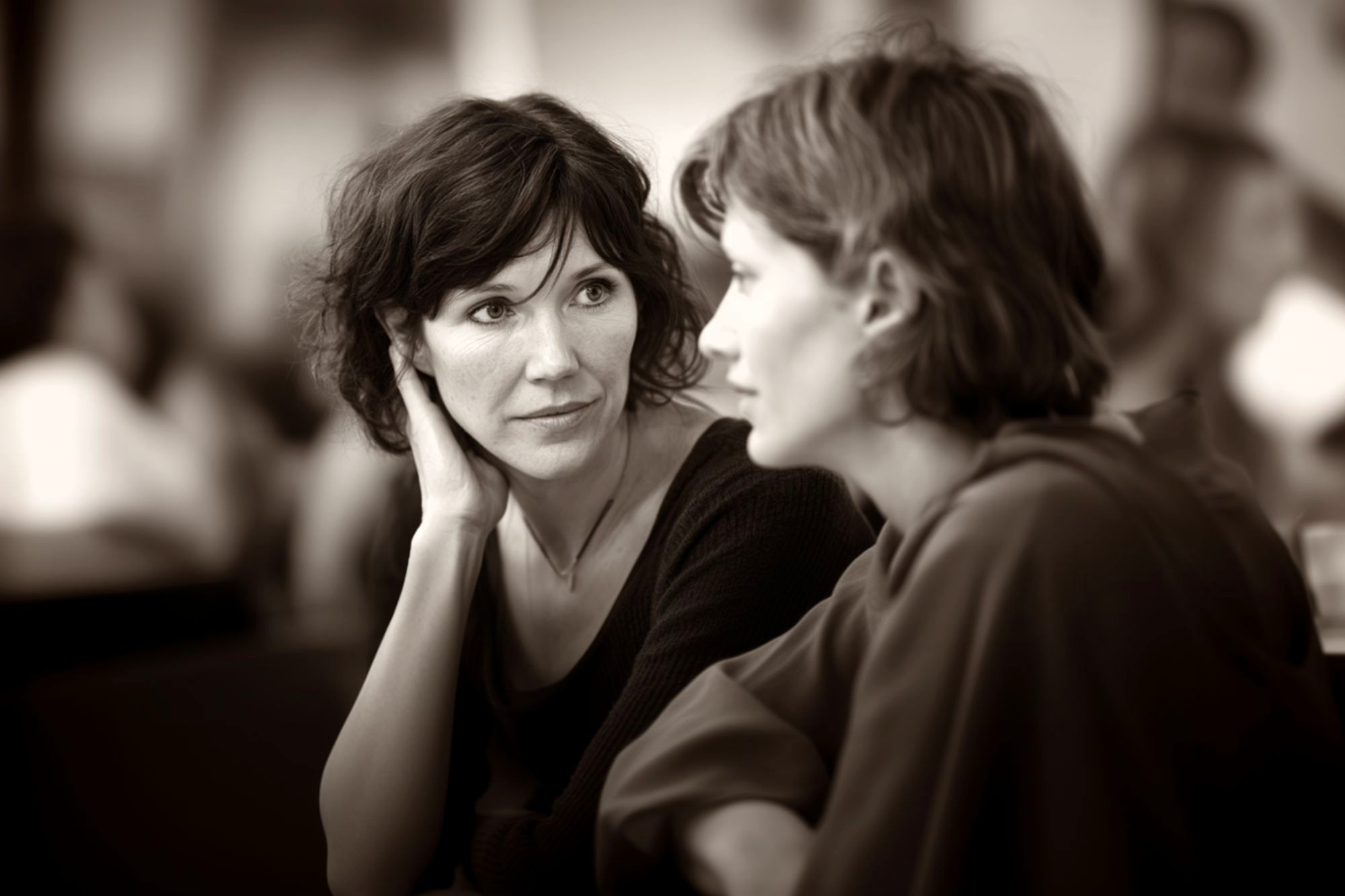 Two women sit close together, having an intimate conversation. One woman listens attentively with her hand to her face, while the other speaks. The background is softly blurred, focusing attention on their expressions.