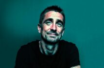 A man with short dark hair, light beard, and mustache wears a black shirt, sitting against a teal background. He is looking slightly up and smiling softly. The image has a digital, artistic filter effect.