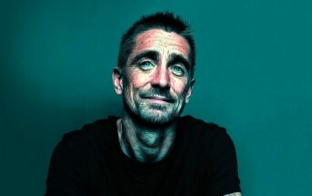 A man with short dark hair, light beard, and mustache wears a black shirt, sitting against a teal background. He is looking slightly up and smiling softly. The image has a digital, artistic filter effect.