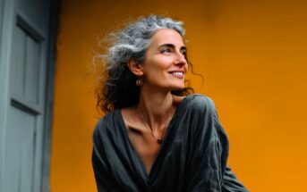 A woman with curly gray hair smiles while looking to the side, wearing a loose gray top and necklace, standing in front of a bright yellow wall.