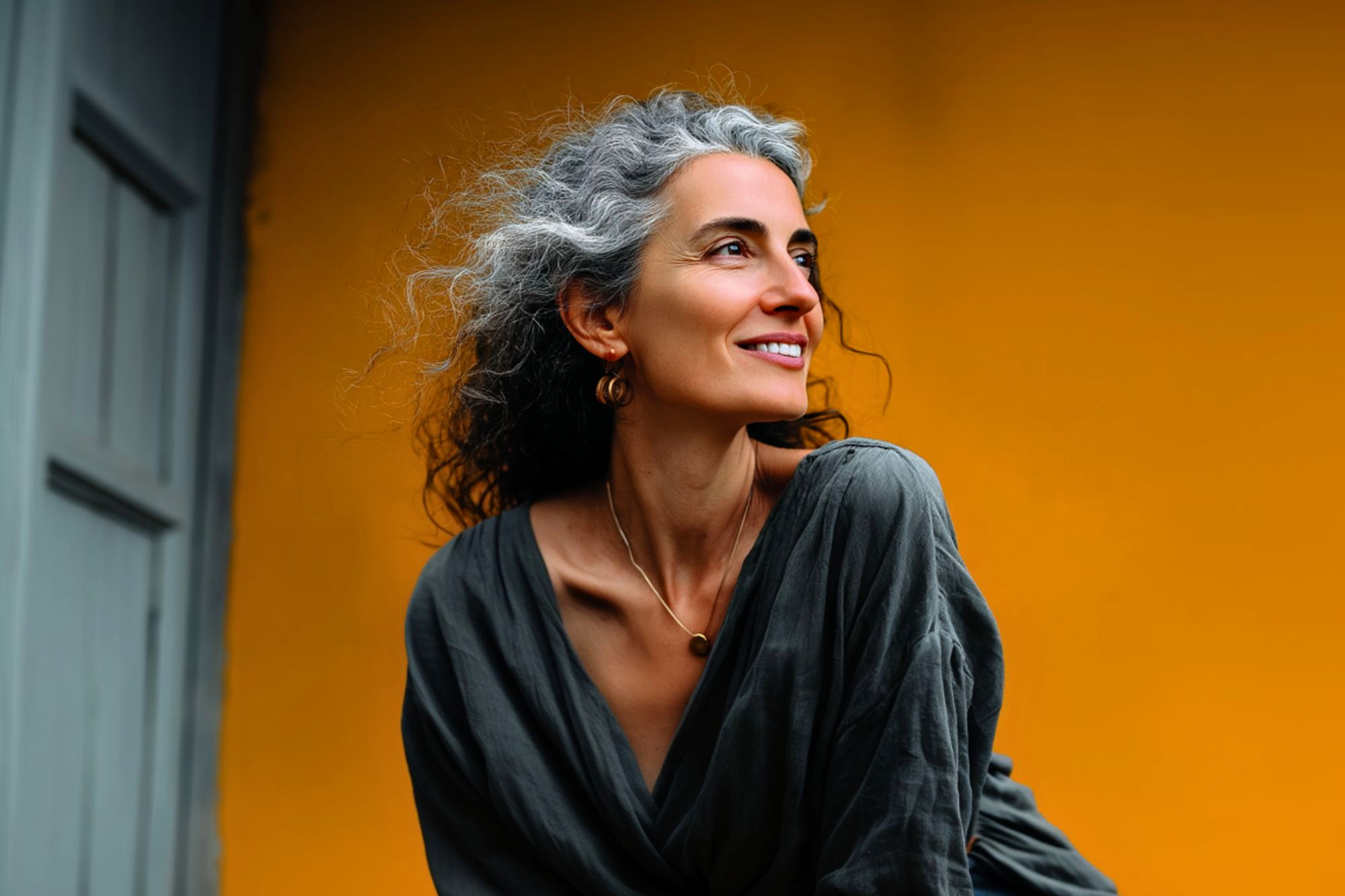 A woman with curly gray hair smiles while looking to the side, wearing a loose gray top and necklace, standing in front of a bright yellow wall.