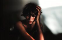 A person with short, tousled hair sits in soft light, resting their head on one hand and looking down with a thoughtful, somber expression. Shadows and highlights create a dramatic, moody atmosphere.