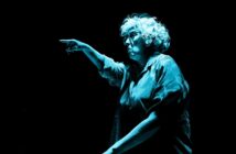 An older person with curly white hair, wearing glasses and a loose shirt, is dramatically pointing forward. The lighting creates a blue tone, highlighting their focused expression against a dark background.
