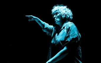 An older person with curly white hair, wearing glasses and a loose shirt, is dramatically pointing forward. The lighting creates a blue tone, highlighting their focused expression against a dark background.