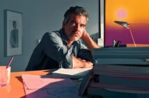 A man with gray hair sits at a desk covered with papers and folders, resting his head on his hand, looking thoughtful. Warm sunlight from a window with a sunset view illuminates the scene.
