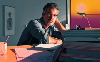 A man with gray hair sits at a desk covered with papers and folders, resting his head on his hand, looking thoughtful. Warm sunlight from a window with a sunset view illuminates the scene.