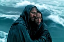 Two people huddled together in rain-soaked cloaks, standing near rough ocean waves, looking weary and solemn as they seek comfort from each other amid the storm.