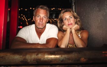 A man and woman sit side by side at a bar. The man looks at the camera while the woman rests her chin on her hands, gazing away. String lights glow softly in the background.