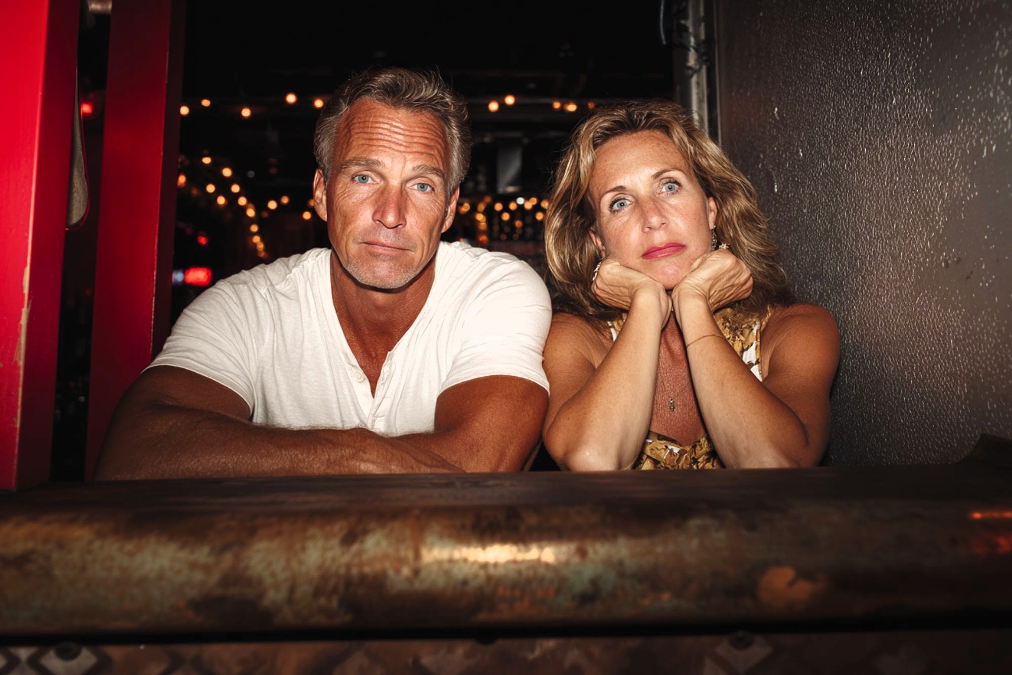 A man and woman sit side by side at a bar. The man looks at the camera while the woman rests her chin on her hands, gazing away. String lights glow softly in the background.