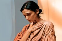 A woman with dark hair pulled back looks down sadly, her hand resting on her chest. She wears a loose, light brown top and stands against a softly lit, warm-toned background.