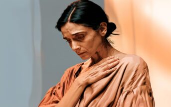 A woman with dark hair pulled back looks down sadly, her hand resting on her chest. She wears a loose, light brown top and stands against a softly lit, warm-toned background.