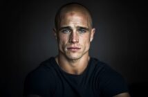 A serious-looking man with a shaved head and blue eyes wears a dark t-shirt, sitting against a dark background. His face shows strong features and subtle wrinkles, with a focused and intense expression.