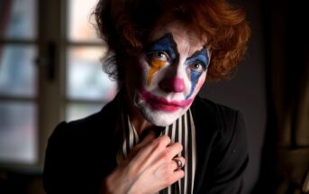 A person with curly hair and colorful clown makeup, wearing a black outfit and striped scarf, looks directly at the camera with a somber expression. A window is blurred in the background.
