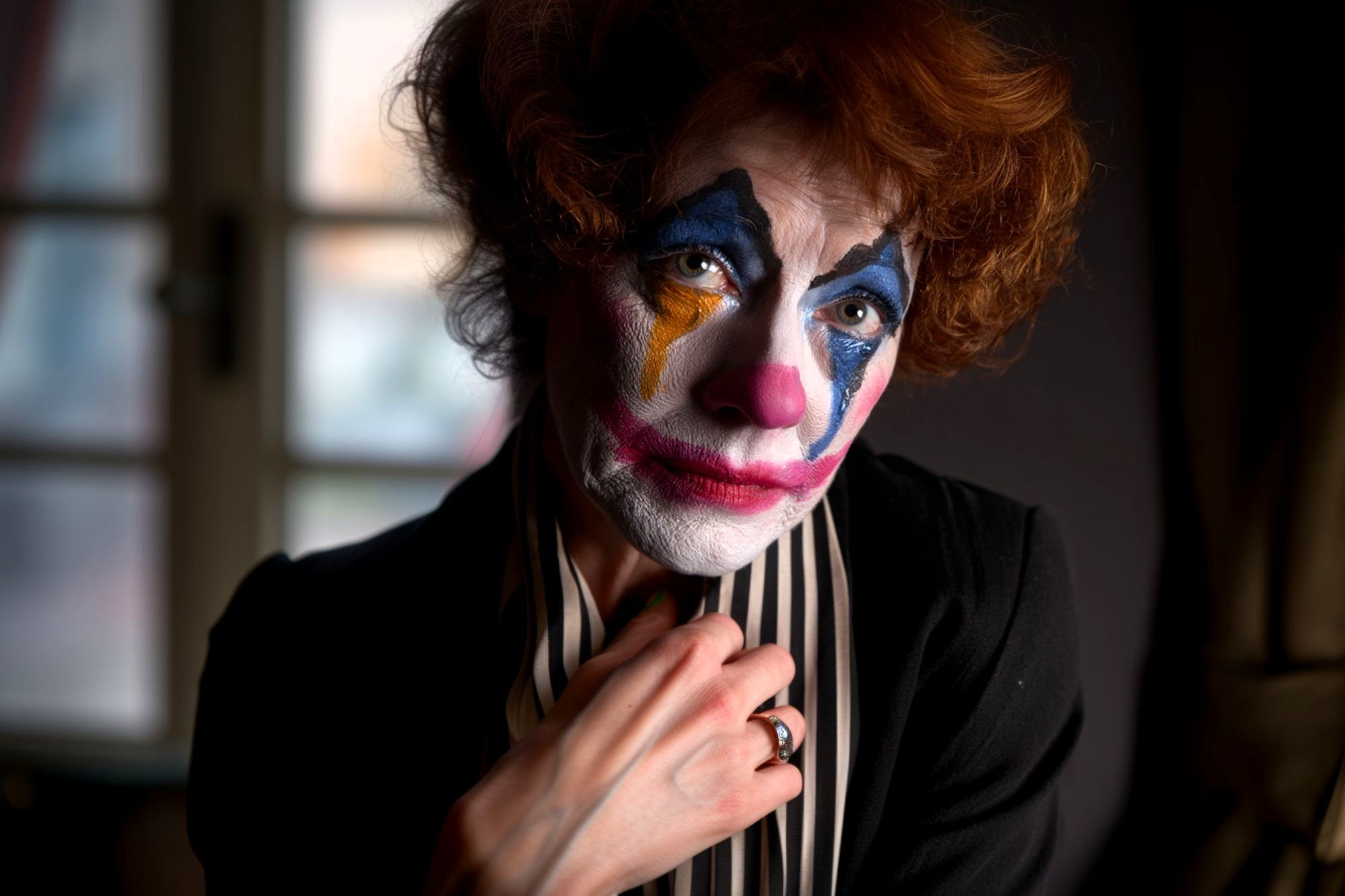 A person with curly hair and colorful clown makeup, wearing a black outfit and striped scarf, looks directly at the camera with a somber expression. A window is blurred in the background.