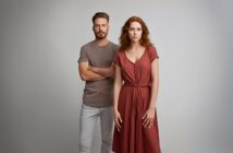 A man in a taupe t-shirt and light pants stands with arms crossed next to a woman in a rust-colored dress, both looking at the camera against a plain light background.