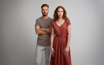 A man in a taupe t-shirt and light pants stands with arms crossed next to a woman in a rust-colored dress, both looking at the camera against a plain light background.