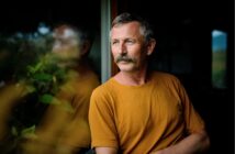 A middle-aged man with a mustache, wearing a mustard yellow shirt, sits indoors near a window, looking thoughtfully outside. Green plants and a soft reflection appear on the glass beside him.