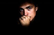 A person with blue eyes looks pensively at the camera, partially covering their mouth with their hand. The face is dramatically lit, surrounded by darkness.