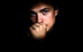A person with blue eyes looks pensively at the camera, partially covering their mouth with their hand. The face is dramatically lit, surrounded by darkness.