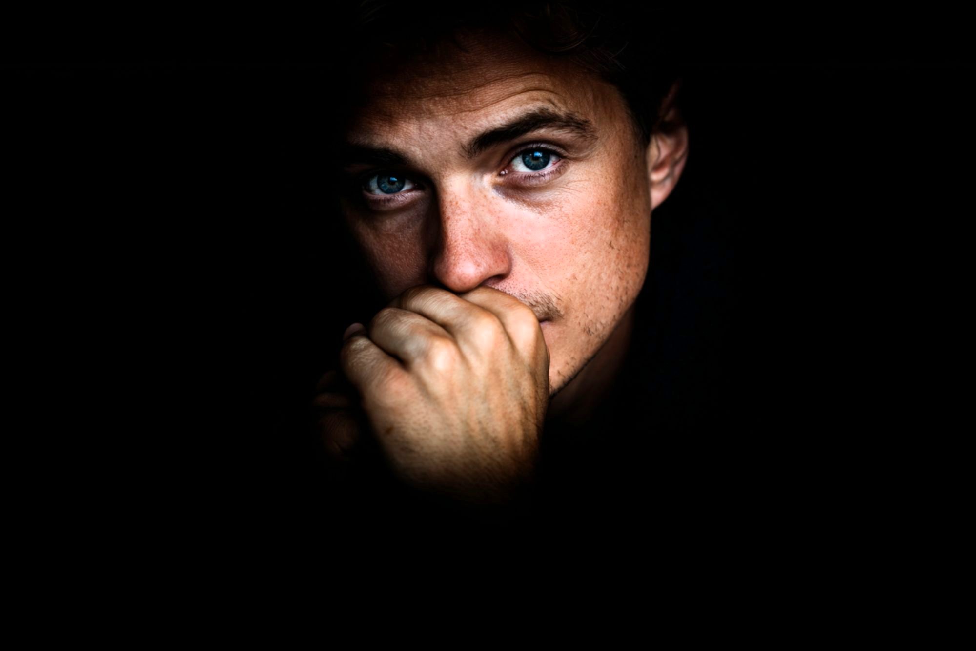 A person with blue eyes looks pensively at the camera, partially covering their mouth with their hand. The face is dramatically lit, surrounded by darkness.