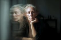 An older woman with short gray hair rests her chin on her hand, staring seriously forward, with a blurred double exposure effect showing her face in profile beside her.