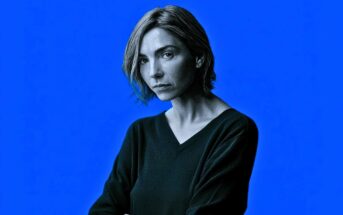 A woman with short, light brown hair and a serious expression looks at the camera. She wears a dark V-neck sweater, standing against a solid bright blue background.