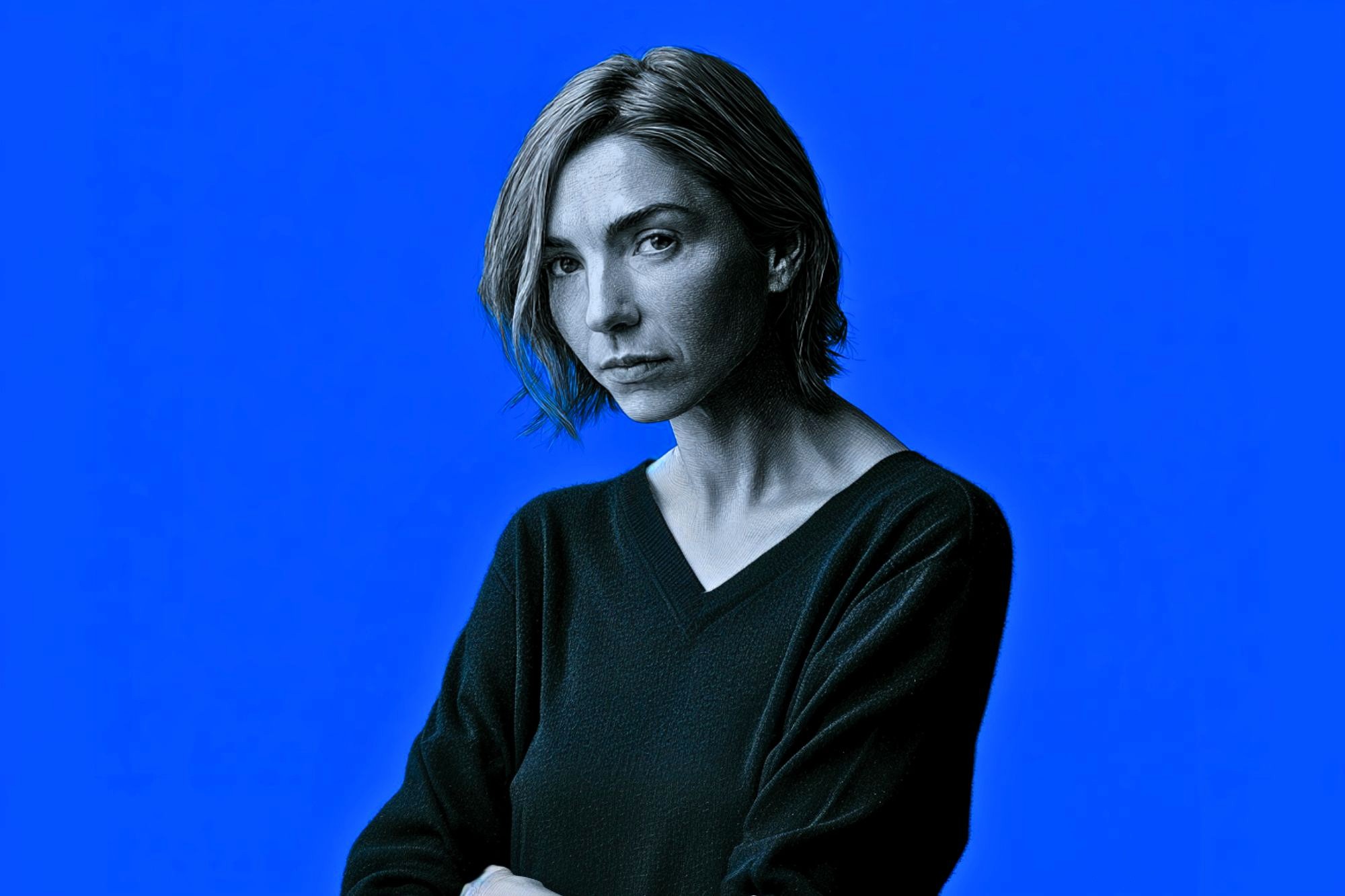 A woman with short, light brown hair and a serious expression looks at the camera. She wears a dark V-neck sweater, standing against a solid bright blue background.