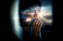 A person with a thoughtful, worried expression looks out a window, resting their hand near their mouth. Light flares and reflections create a dreamy, introspective mood.