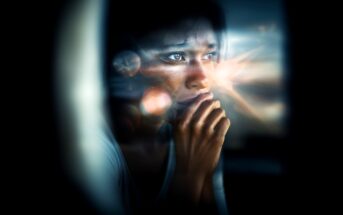 A person with a thoughtful, worried expression looks out a window, resting their hand near their mouth. Light flares and reflections create a dreamy, introspective mood.