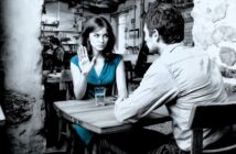 A woman in a blue dress raises her hand in refusal while sitting across from a man at a cafe table. Both look serious, and there are glasses of water on the table. The setting is rustic with stone walls.