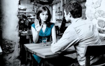 A woman in a blue dress raises her hand in refusal while sitting across from a man at a cafe table. Both look serious, and there are glasses of water on the table. The setting is rustic with stone walls.