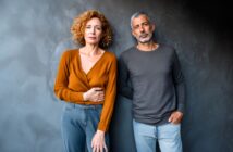 A woman with curly hair in an orange top and a man with short gray hair in a gray shirt stand next to each other against a dark textured wall, both looking seriously at the camera.