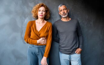 A woman with curly hair in an orange top and a man with short gray hair in a gray shirt stand next to each other against a dark textured wall, both looking seriously at the camera.