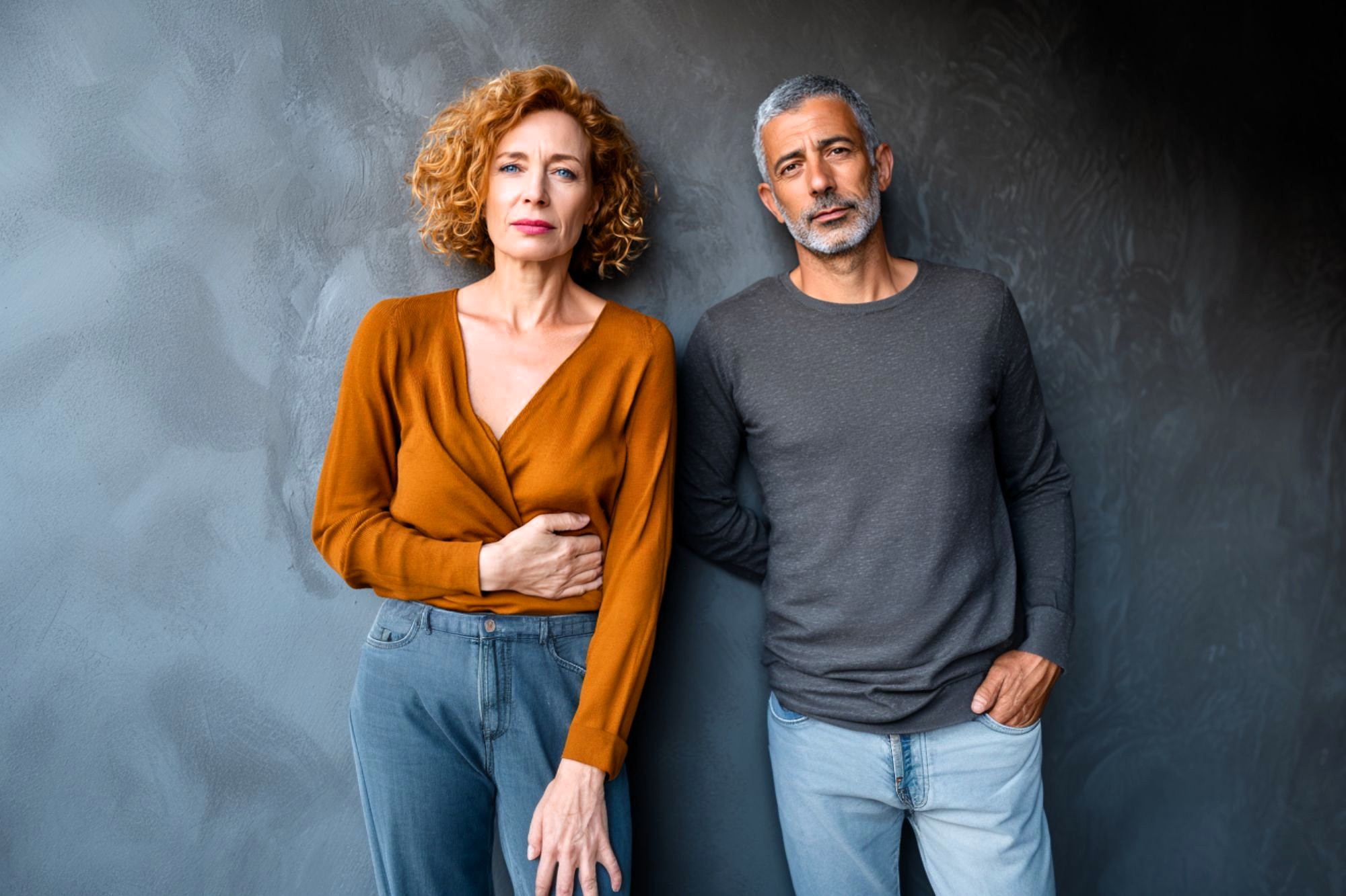 A woman with curly hair in an orange top and a man with short gray hair in a gray shirt stand next to each other against a dark textured wall, both looking seriously at the camera.